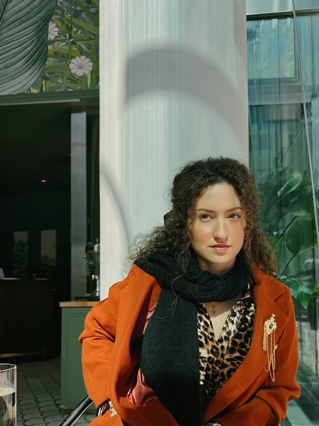 Portrait of Autumn Lundberg in a rust-orange coat and black scarf, seated in sunlight in front of a white column.