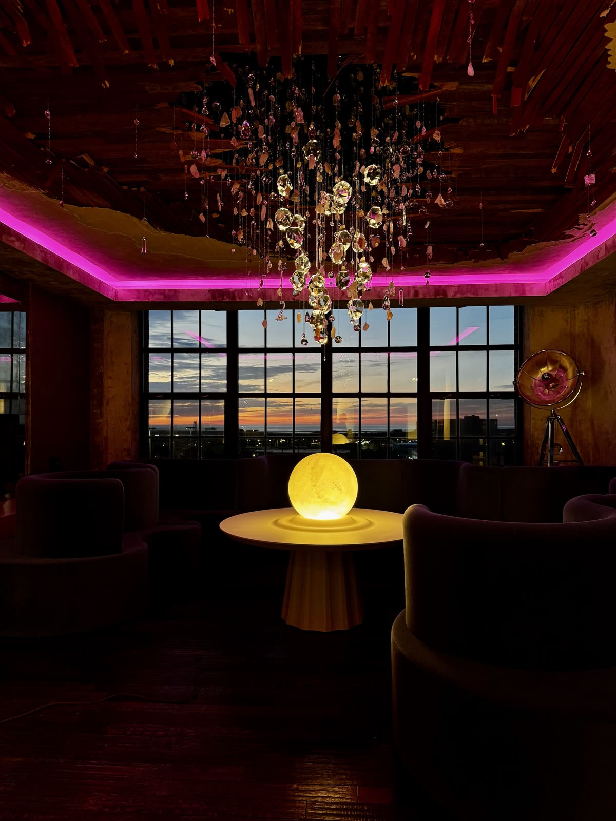 The installation at dusk: the crystal cascade glowing gold beneath the torn lath ceiling, a pink light cove around the room, a glowing sphere on a table and sunset through the windows.