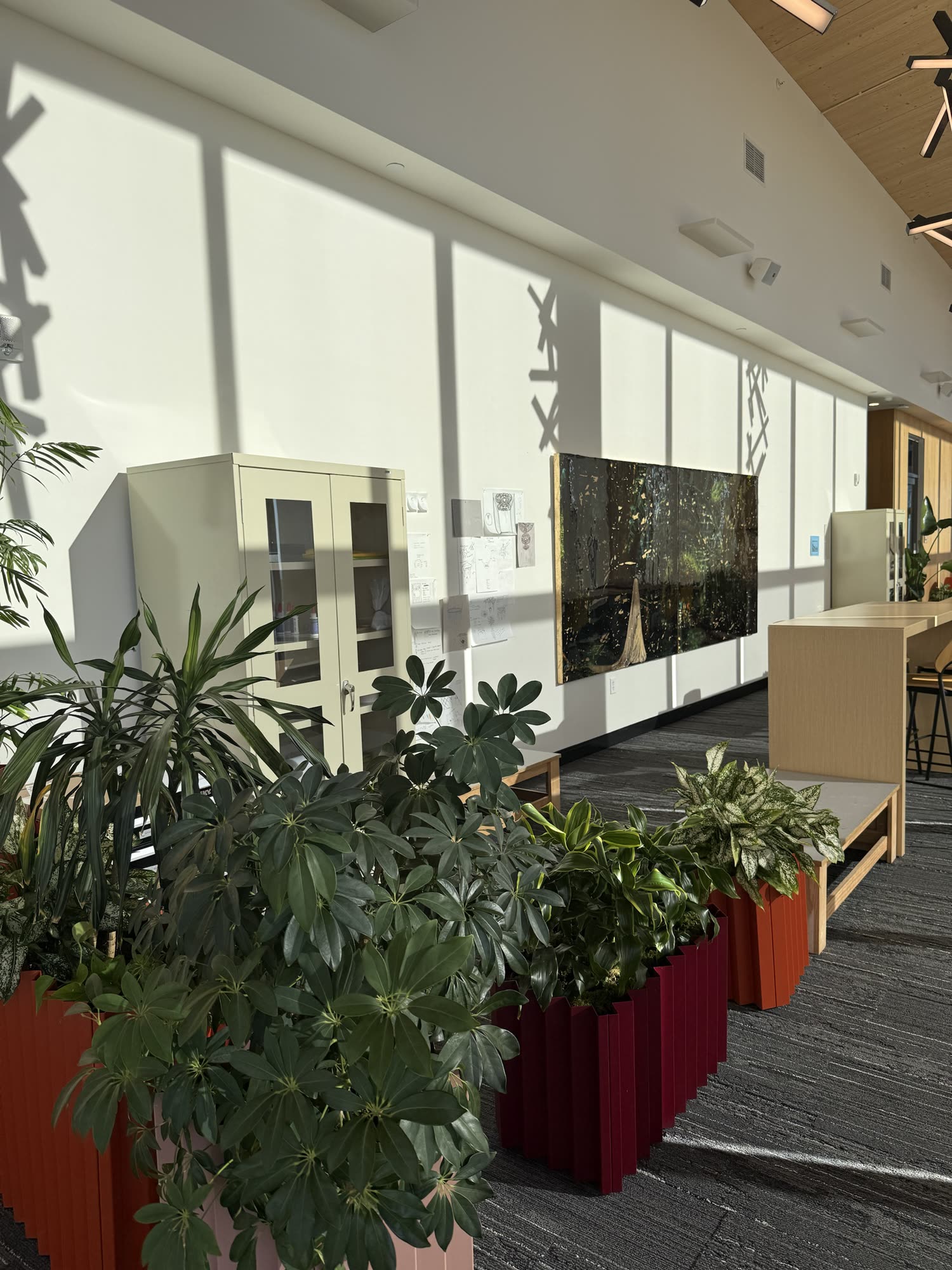 The artist in residence studio: a sunlit white wall with a large dark painting, pinned sketches and a cabinet, under a timber ceiling, with planters in the foreground.