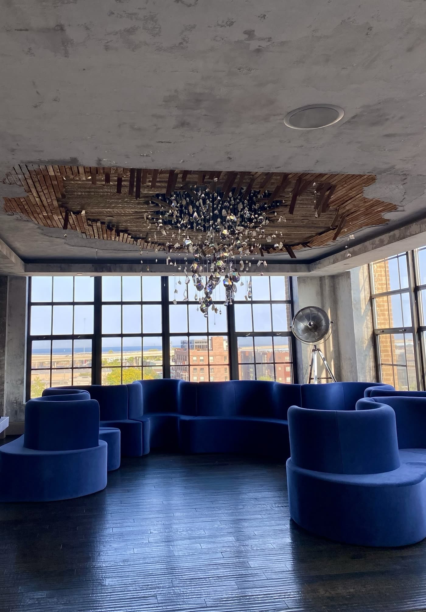 The finished installation in a historic warehouse loft: a jagged opening of wood lath in the ceiling, crystals cascading toward a curved cobalt velvet sofa and tall industrial windows.